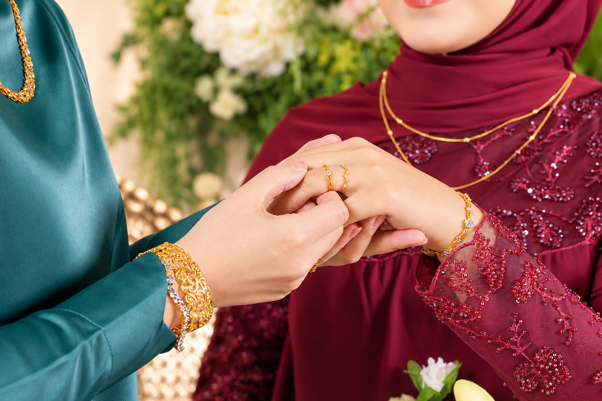 A close-up lifestyle shot of a gold ring being placed on a woman's finger. The woman is wearing a deep burgundy lace hijab and matching dress, accessorized with gold necklaces and bracelets, while another person wears a teal satin top and a gold bangle.